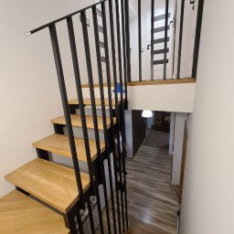 DREWBUDSYSTEM PROSTA SPÓŁKA AKCYJNA - Drewniane schody z czarną metalową balustradą w nowoczesnym wnętrzu, widok z góry schodów na dół, z widocznym przejściem do innego pomieszczenia.