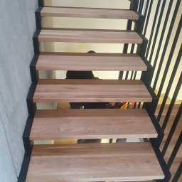 DREWBUDSYSTEM PROSTA SPÓŁKA AKCYJNA - Nowoczesne schody z drewnianymi stopniami i czarną metalową konstrukcją, widok z dołu, minimalistyczny design.