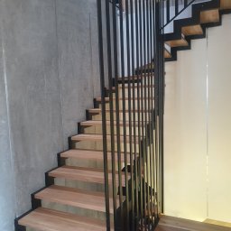 DREWBUDSYSTEM PROSTA SPÓŁKA AKCYJNA - Nowoczesne schody drewniane z czarną metalową balustradą w minimalistycznym wnętrzu z betonową ścianą.