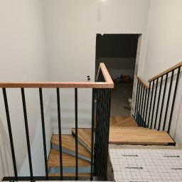 DREWBUDSYSTEM PROSTA SPÓŁKA AKCYJNA - Nowe schody z drewnianymi stopniami i czarnymi metalowymi balustradami w trakcie montażu, zabezpieczone folią, widok z góry na dół.