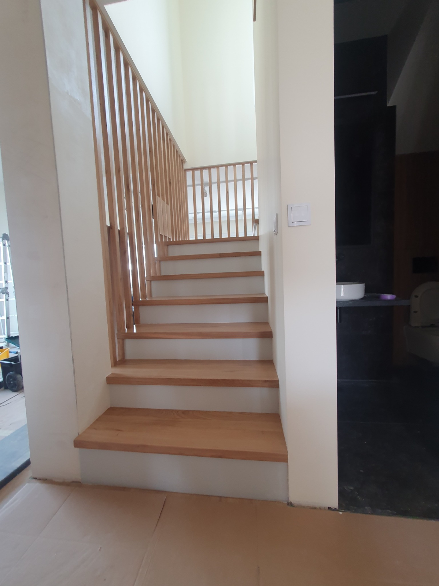 Jasne drewniane schody z barierką z pionowych szczebli w nowym domu, widok z dołu z zabezpieczoną podłogą i fragmentem łazienki w tle.
