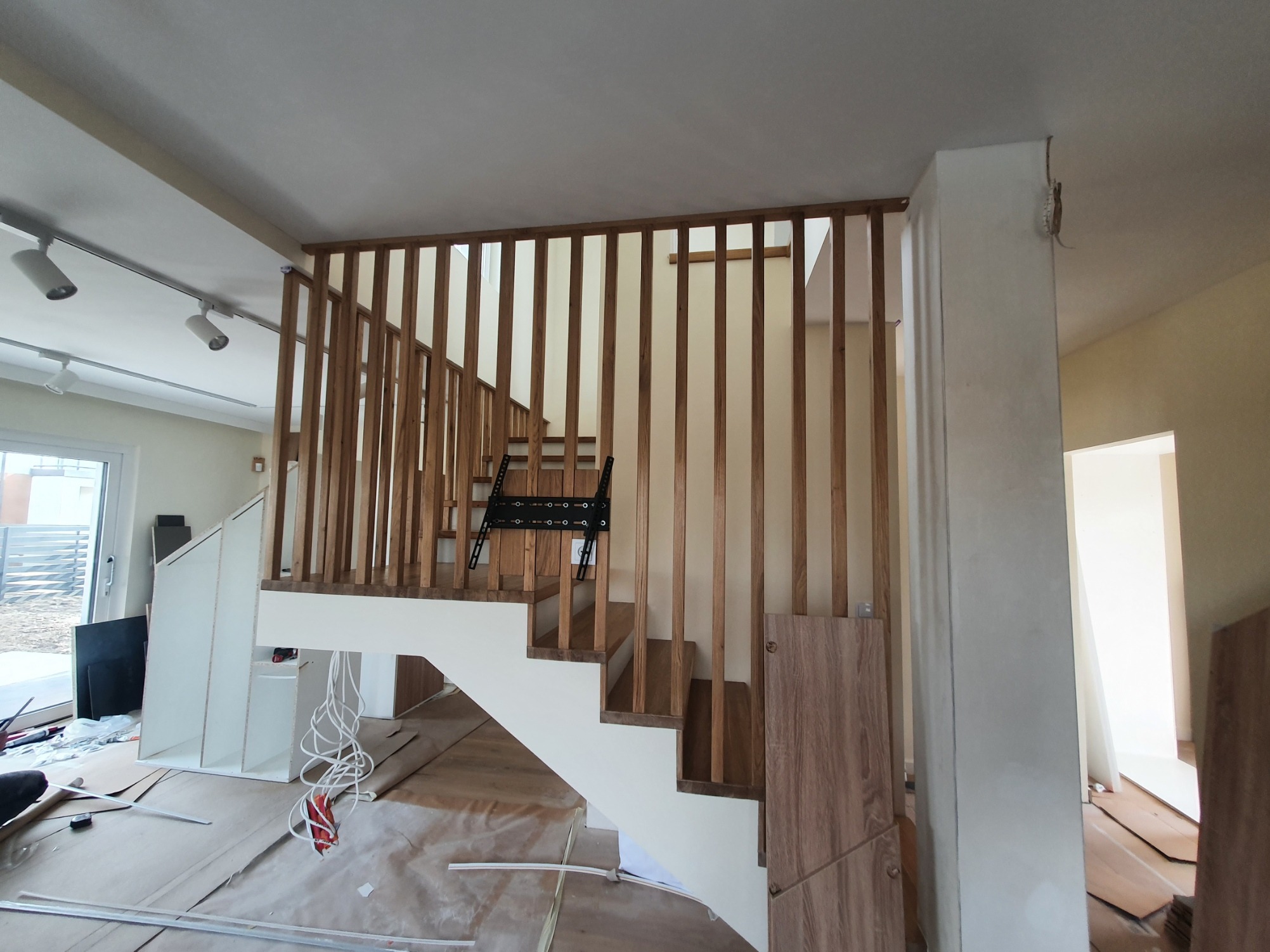 Montaż schodów z drewnianą balustradą w trakcie realizacji. Widoczne zabezpieczenia podłogi, elementy montażowe i wiszący uchwyt na telewizor.