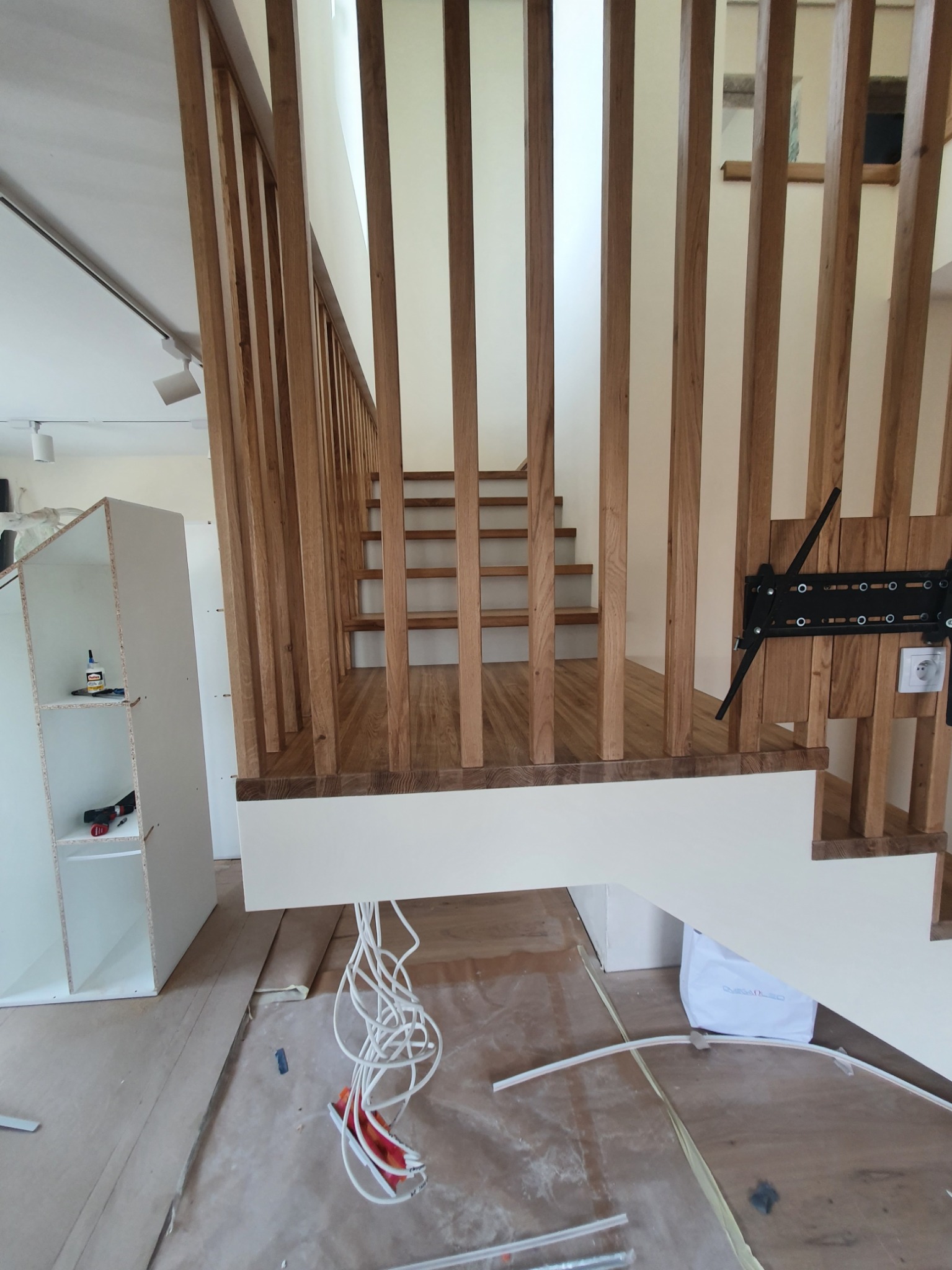 Wnętrze w trakcie remontu z widocznymi drewnianymi schodami z pionowymi balustradami i wystającymi kablami elektrycznymi oraz elementami mebli do złożenia w tle.
