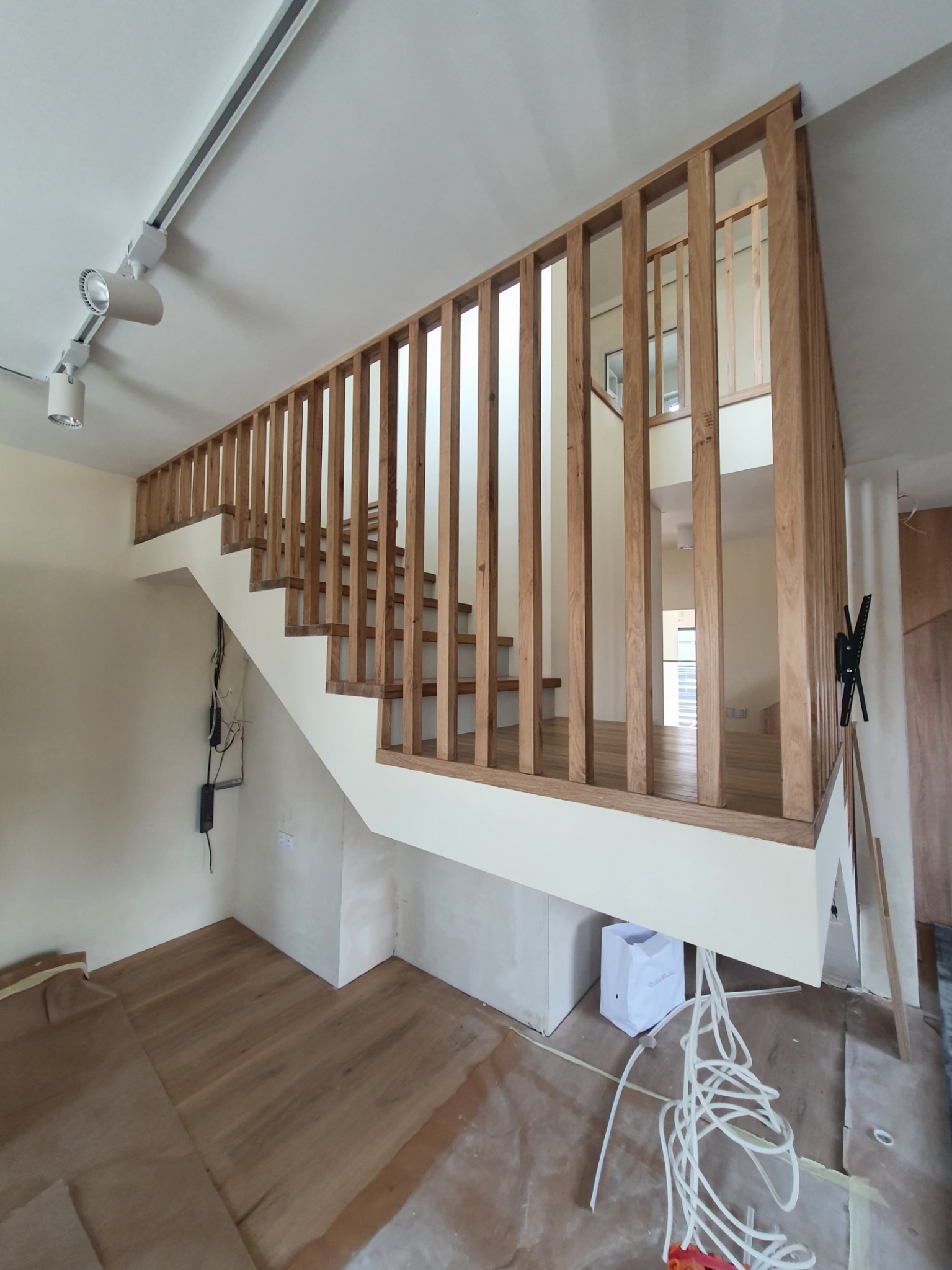 Wykończone drewniane schody z jasnymi, pionowymi balustradami w trakcie prac wykończeniowych, widoczne kable i zabezpieczona podłoga.