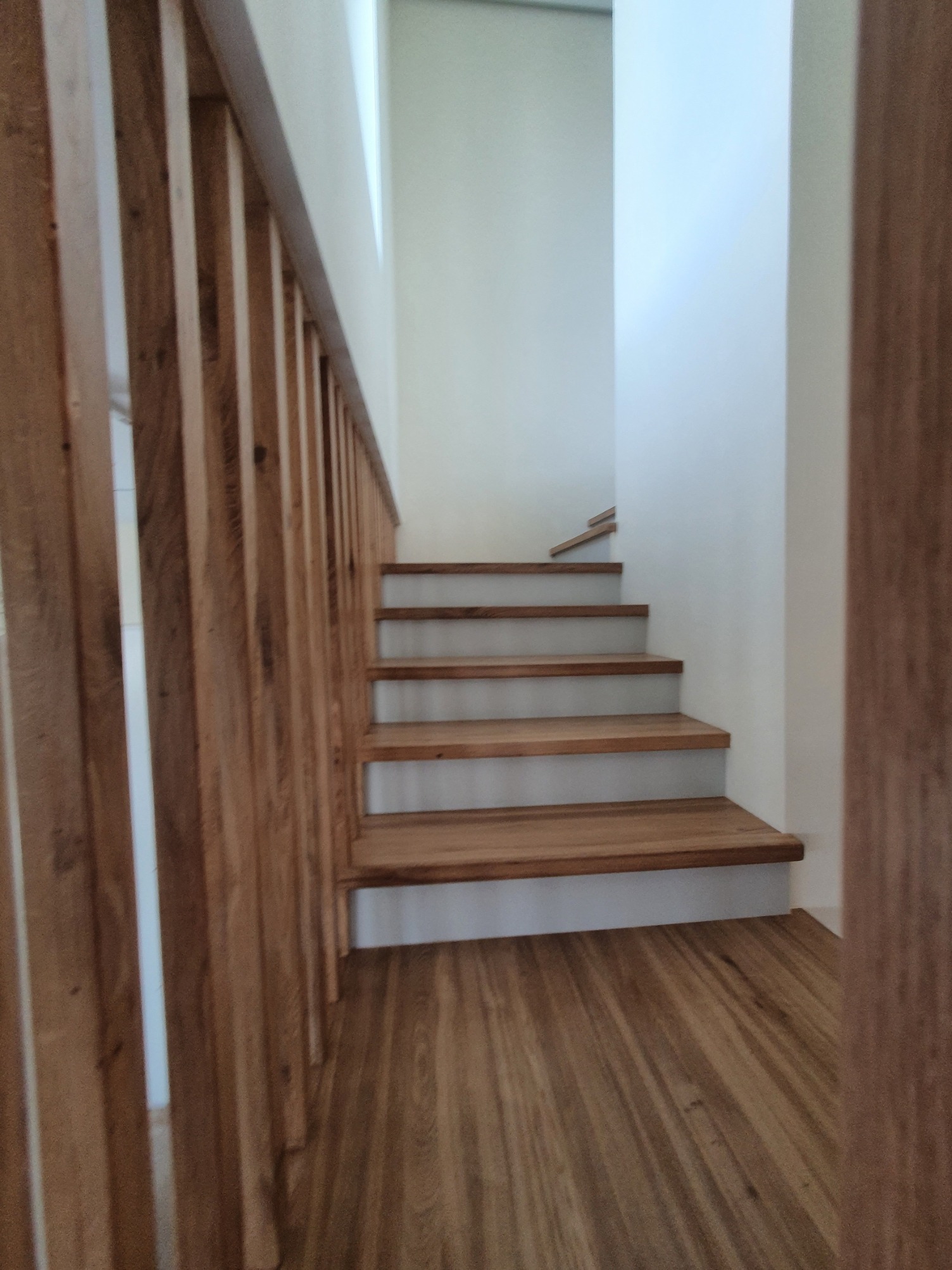 Proste, drewniane schody z jasnymi stopniami i minimalistyczną balustradą z pionowych listew, prowadzące na wyższą kondygnację w jasnym wnętrzu.