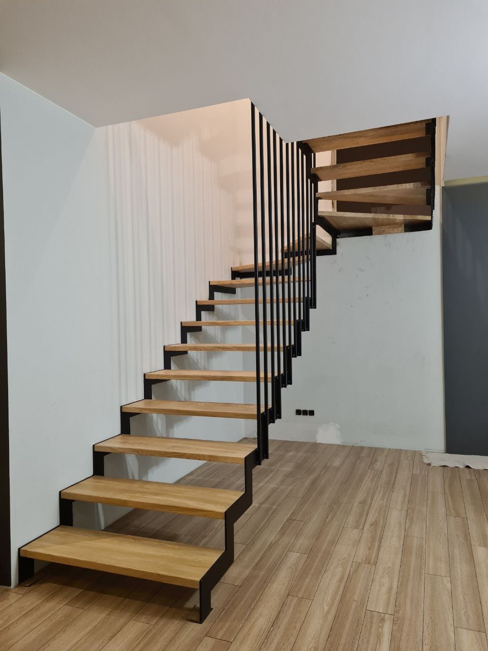 Nowoczesne schody z drewnianymi stopniami i czarną, minimalistyczną metalową konstrukcją, widoczne na tle jasnych ścian i drewnianej podłogi.