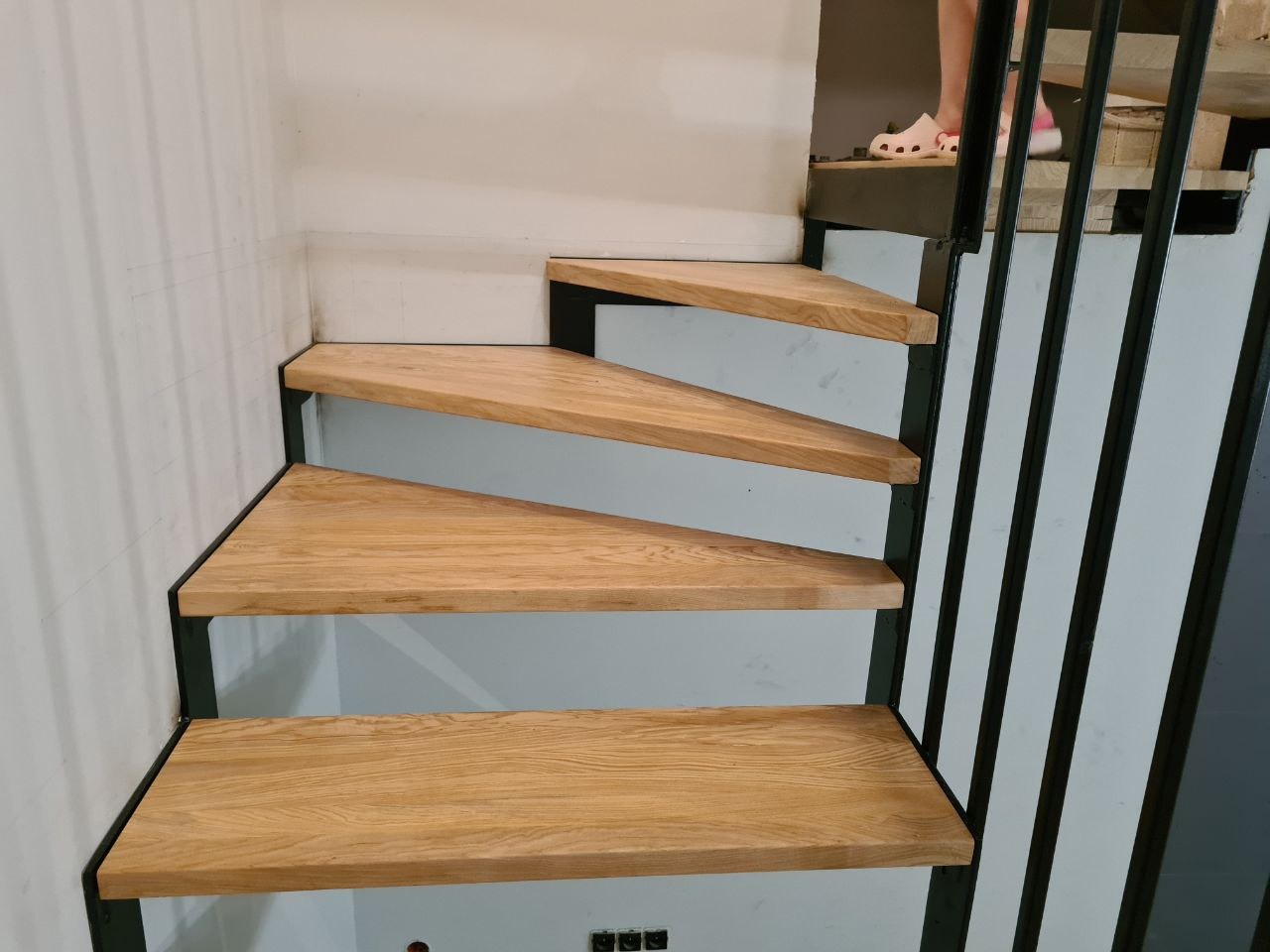 Bieg schodów z drewnianymi stopniami i metalową balustradą, widoczne z dołu, na górze stopy w klapkach.