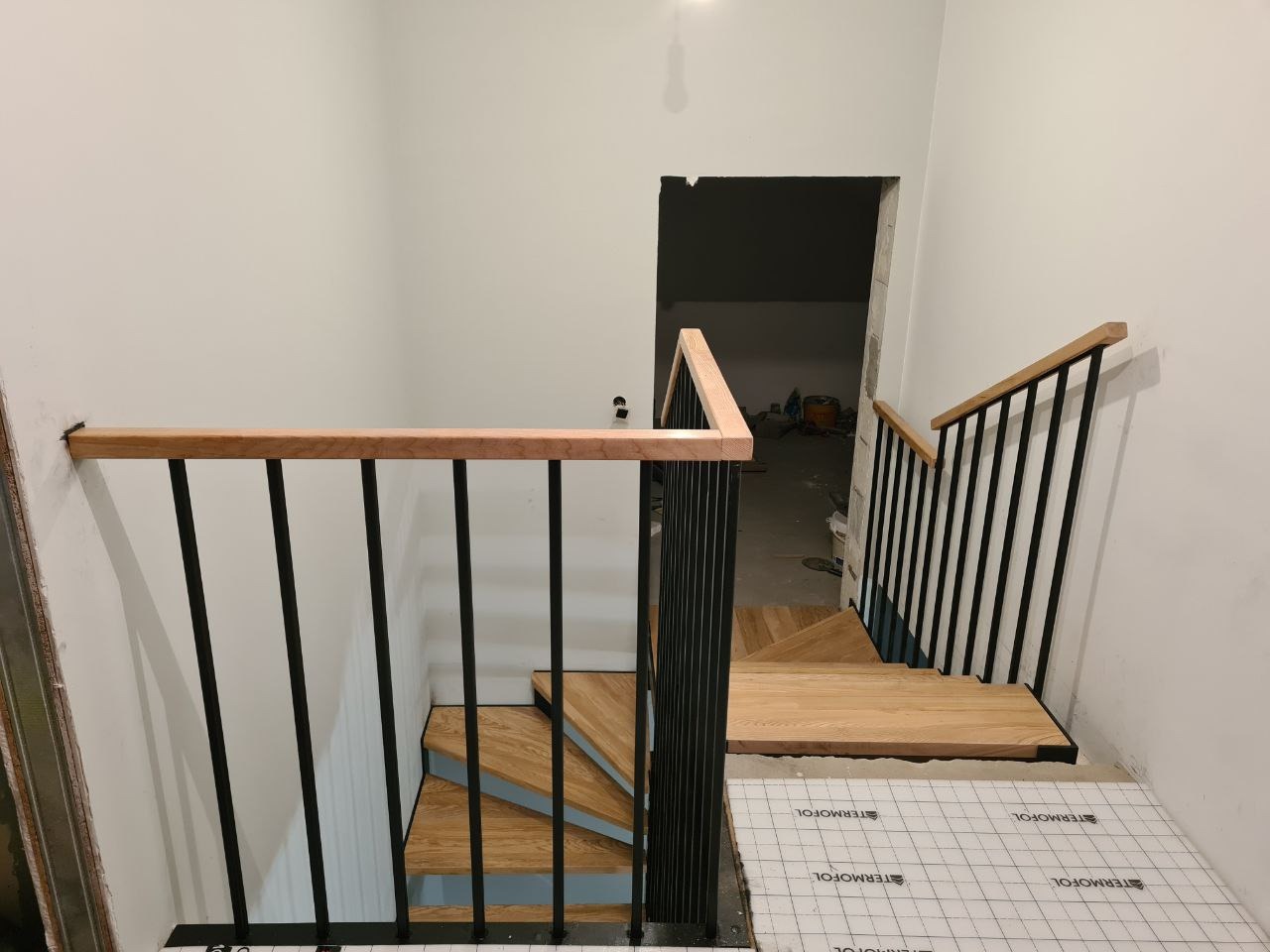 Nowe schody z drewnianymi stopniami i czarnymi metalowymi balustradami w trakcie montażu, zabezpieczone folią, widok z góry na dół.