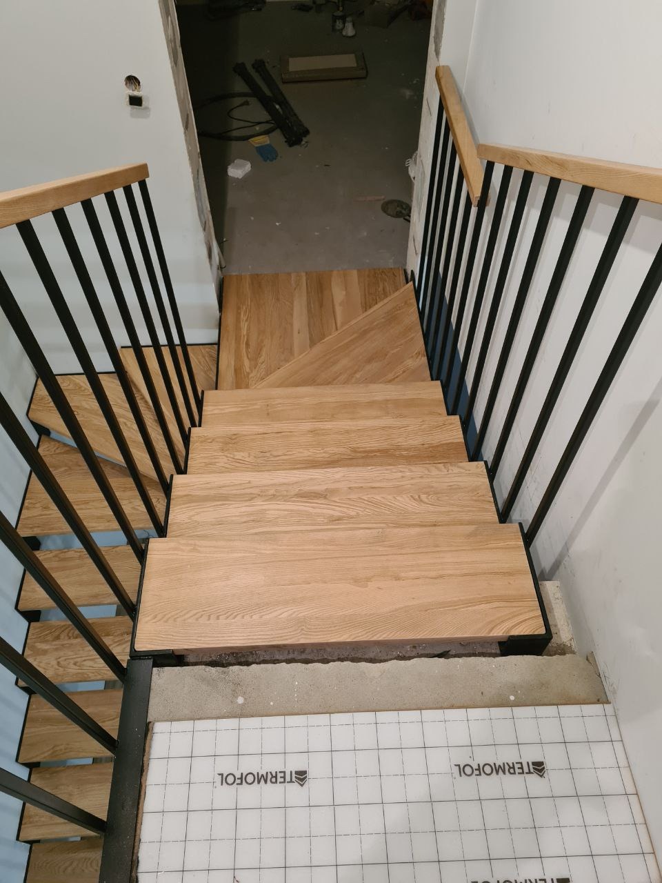 Widok z góry na drewniane stopnie schodów z czarnymi metalowymi balustradami, w trakcie montażu, z widocznym ociepleniem podłoża.