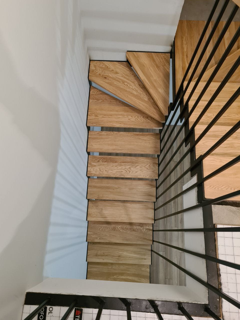 Widok z góry na drewniane schody z czarnymi metalowymi barierkami, stopnie z jasnego drewna o widocznej strukturze, białe ściany i podłoga z białych płytek.