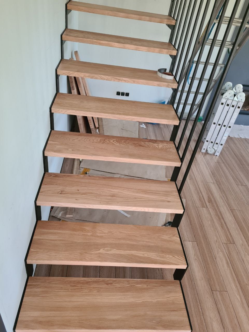 Nowoczesne schody z drewnianymi stopniami i czarną metalową konstrukcją, widok z dołu, w tle drabina aluminiowa.