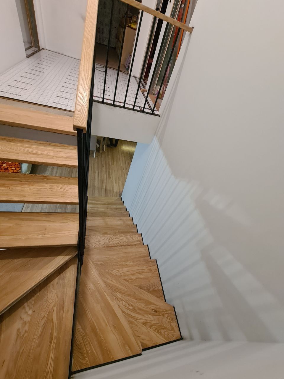 Klatka schodowa z drewnianymi stopniami z wyraźnym usłojeniem, czarnymi krawędziami i metalową balustradą z drewnianym poręczą, widok z góry na dół z perspektywy osoby stojącej na górze schodów.