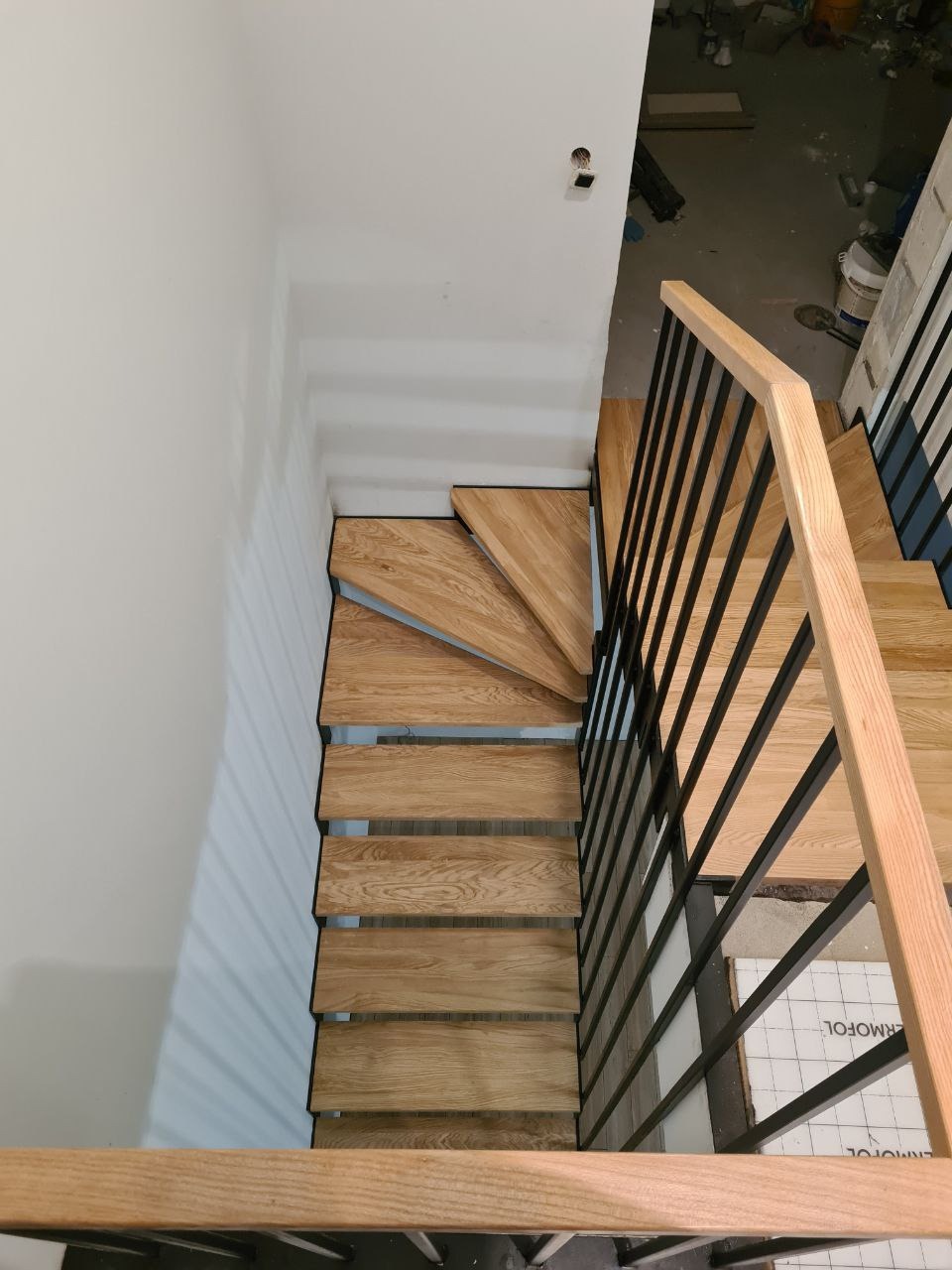Widok z góry na drewniane schody z czarną metalową balustradą, z widocznymi niedoskonałościami wykończenia wnętrza.