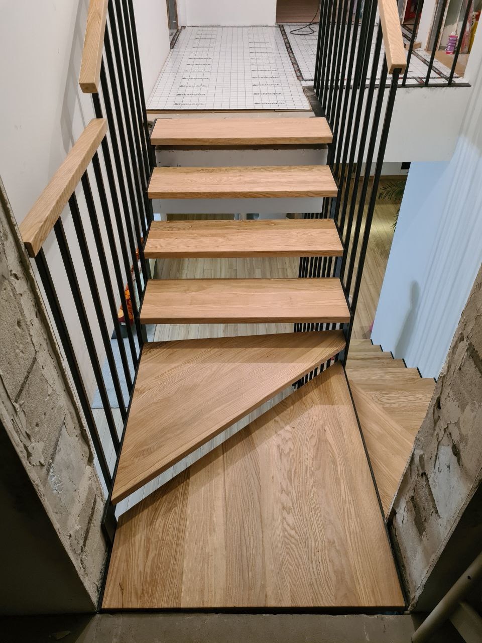 Nowoczesne schody z drewnianymi stopniami i czarnymi metalowymi poręczami, widok z góry na stopnie o nieregularnym kształcie, surowy beton na ścianach.
