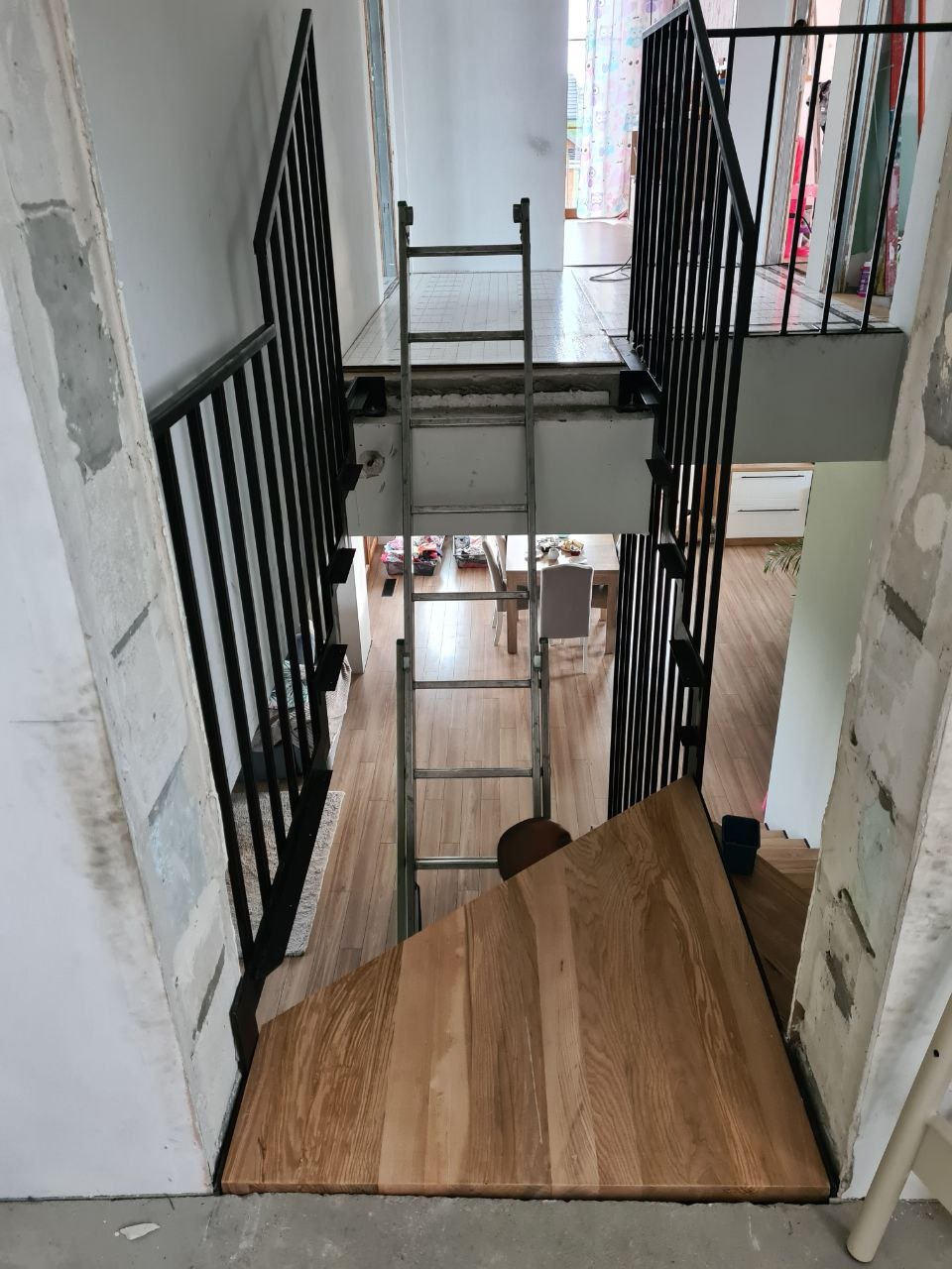 Widok z góry na częściowo wykończone schody z drewnianymi stopniami i metalową balustradą, z drabiną opartą o antresolę i widocznym fragmentem pomieszczenia poniżej.