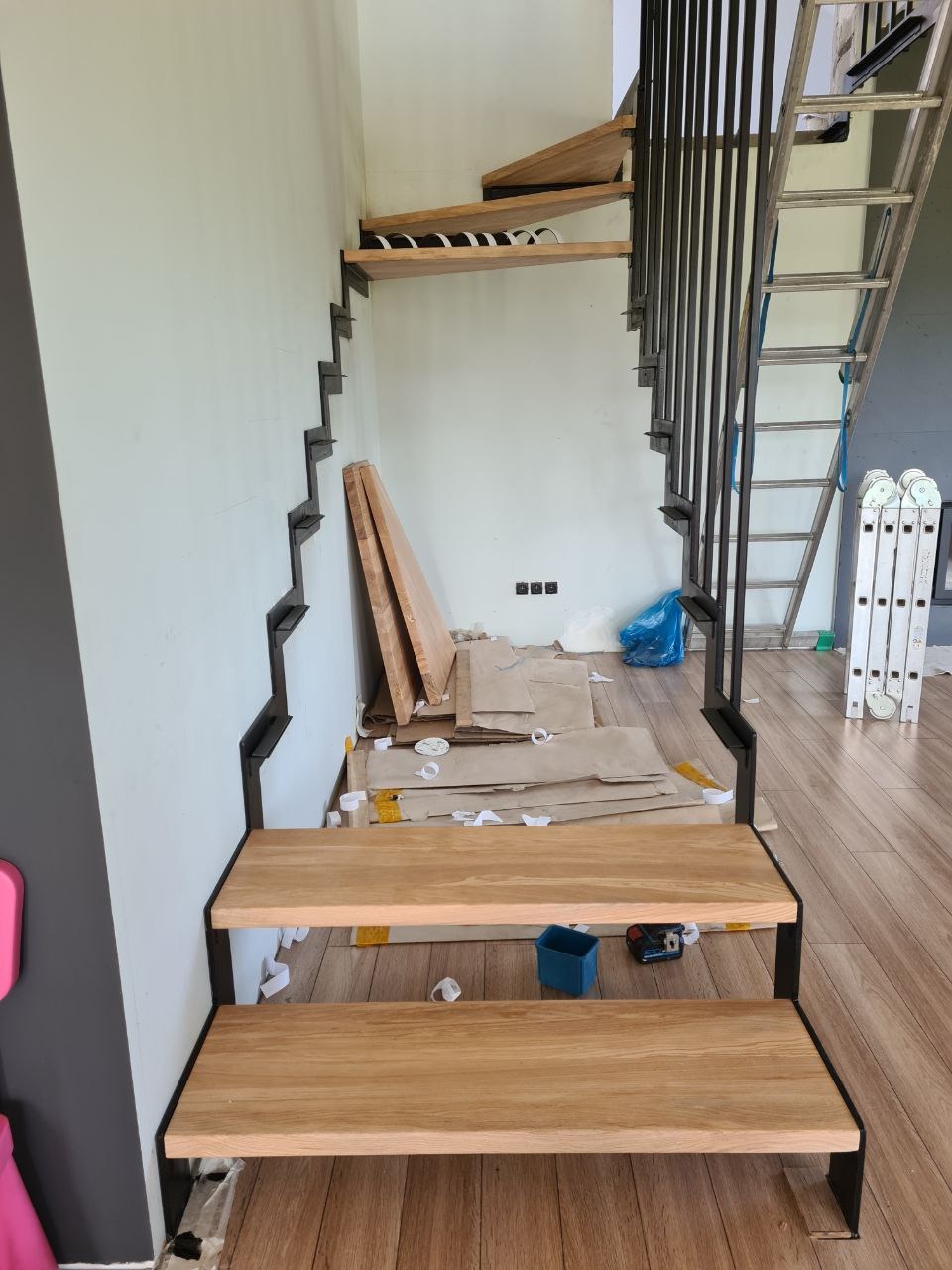 Montaż ażurowych schodów z drewnianymi stopniami i metalową konstrukcją, zabezpieczone kartonem, widoczne narzędzia i drabina.