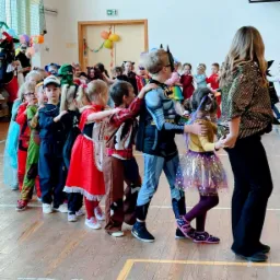 Dzieci w kostiumach, m.in. Batman, ustawione w rzędzie podczas zabawy z animatorką w błyszczącej bluzce na sali gimnastycznej.