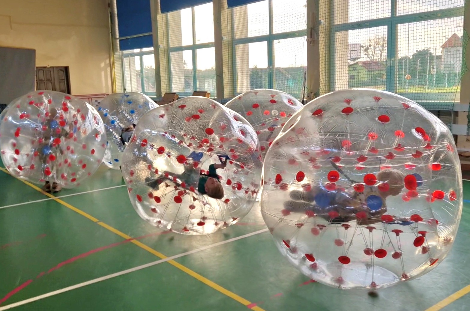 Dzieci w przezroczystych kulach z czerwonymi i niebieskimi elementami, zabawa na zielonej hali sportowej z oknami w tle.
