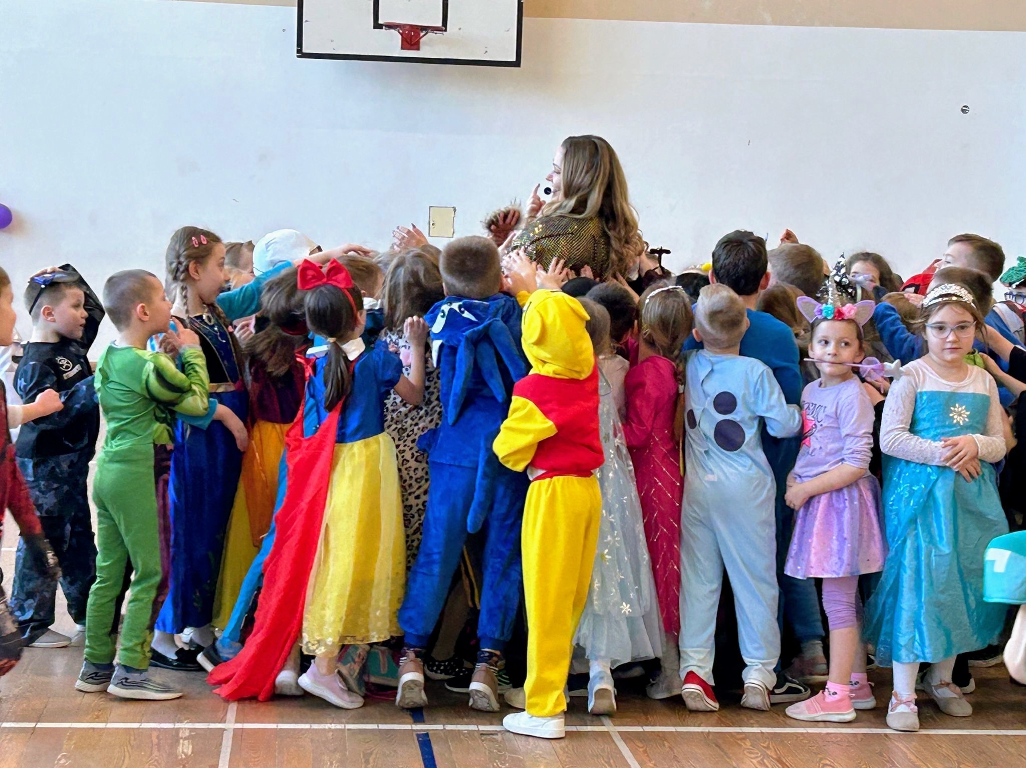 Grupa dzieci w kostiumach postaci z bajek otacza kobietę z mikrofonem na tle białej ściany w sali gimnastycznej, widoczny kosz do koszykówki.