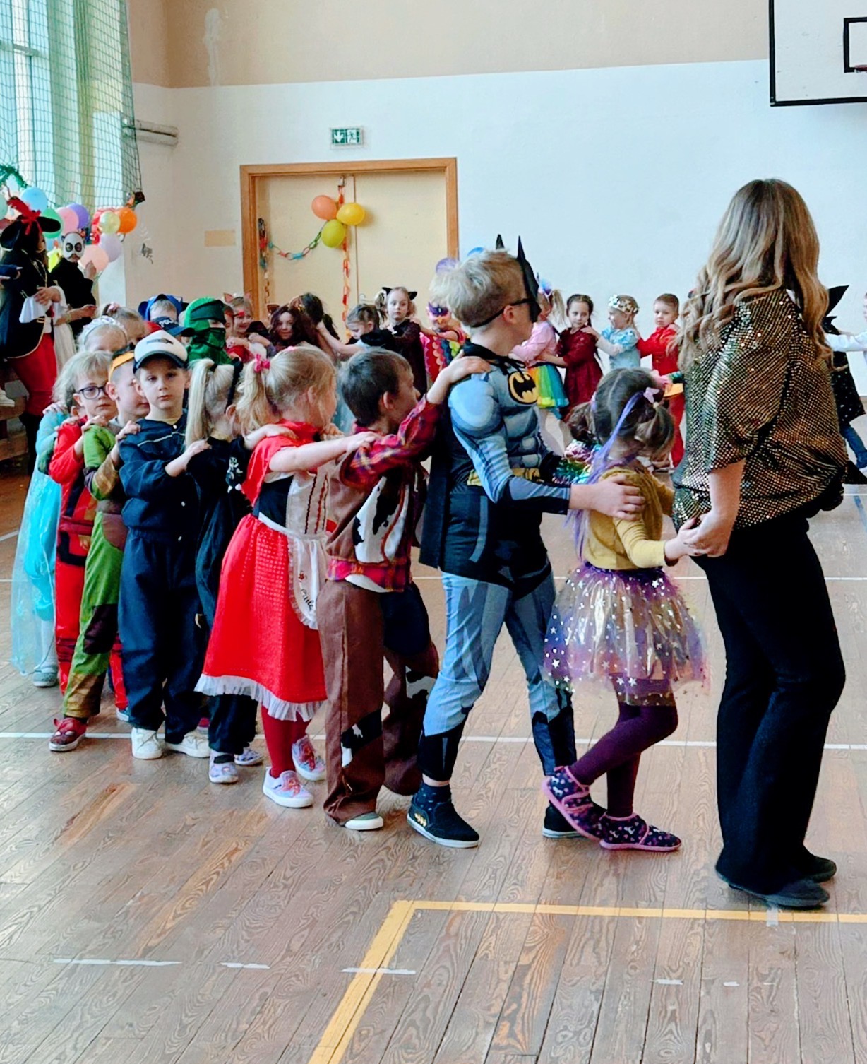 Dzieci w kostiumach, m.in. Batman, ustawione w rzędzie podczas zabawy z animatorką w błyszczącej bluzce na sali gimnastycznej.