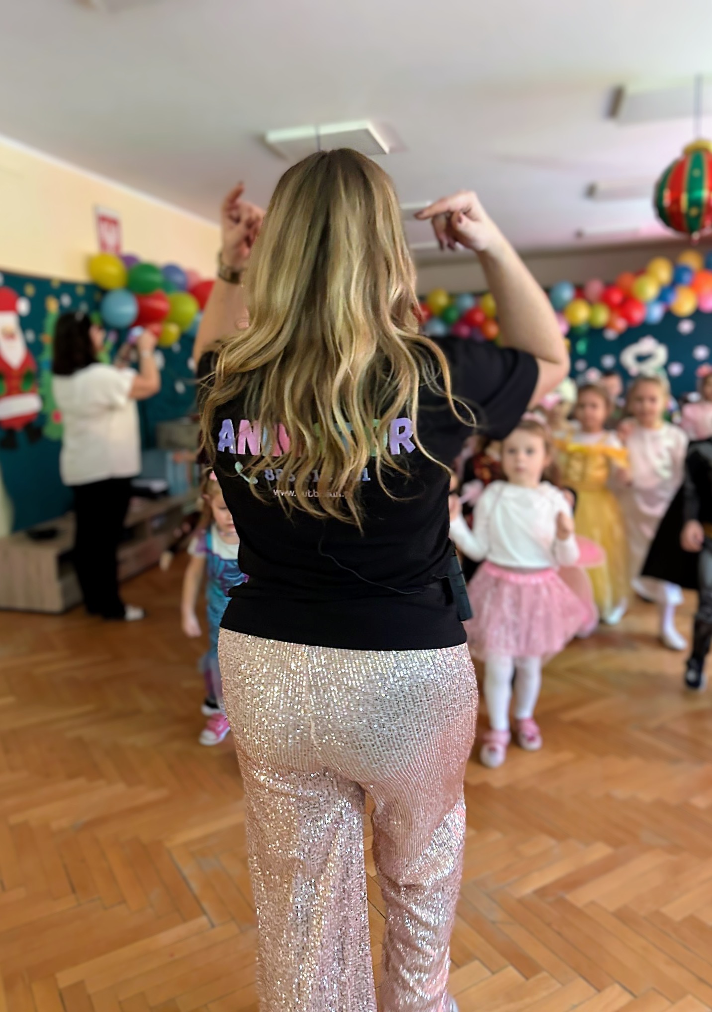 Animator z blond włosami, w czarnej koszulce z napisem i błyszczących spodniach, prowadzi zabawę dla grupy dzieci w strojach karnawałowych w udekorowanym pomieszczeniu.