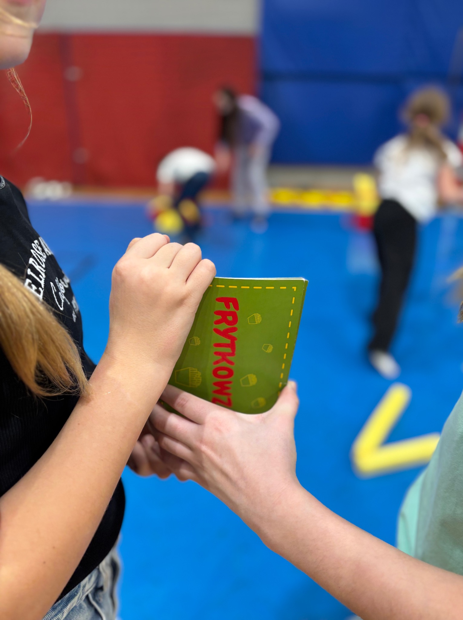 Dziecko trzyma zieloną książeczkę z napisem 'Frytkowy Łowca' podczas zabawy z animatorem w tle na niebieskiej sali gimnastycznej.