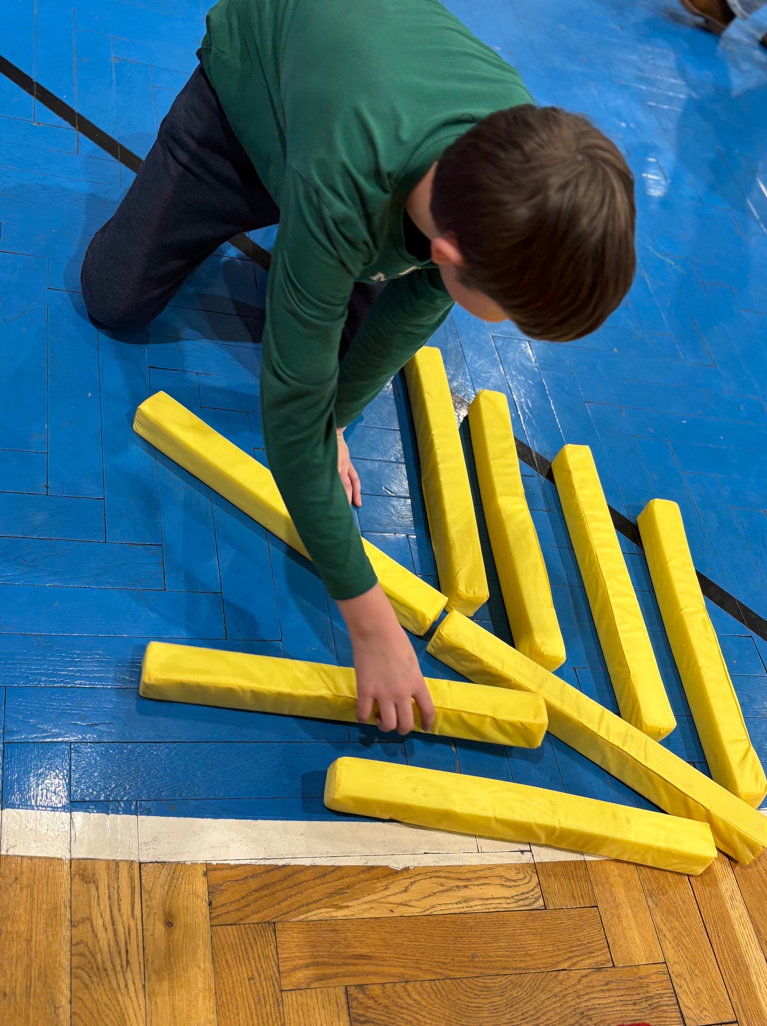 Chłopiec w zielonej bluzce układa żółte, piankowe klocki na niebiesko-brązowej podłodze sali gimnastycznej.