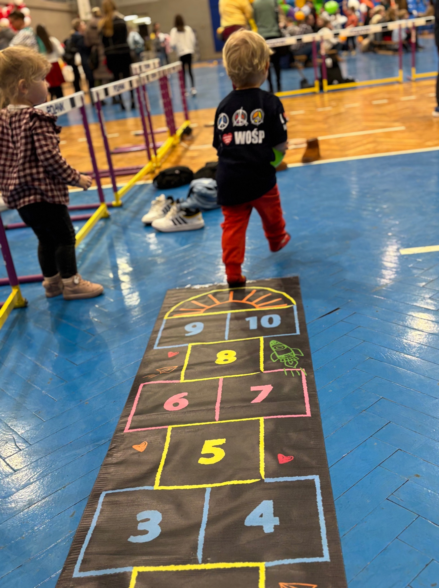 Dziecko w czerwonych spodniach skacze po planszy do gry w klasy na tle sali gimnastycznej z przeszkodami i innymi dziećmi.