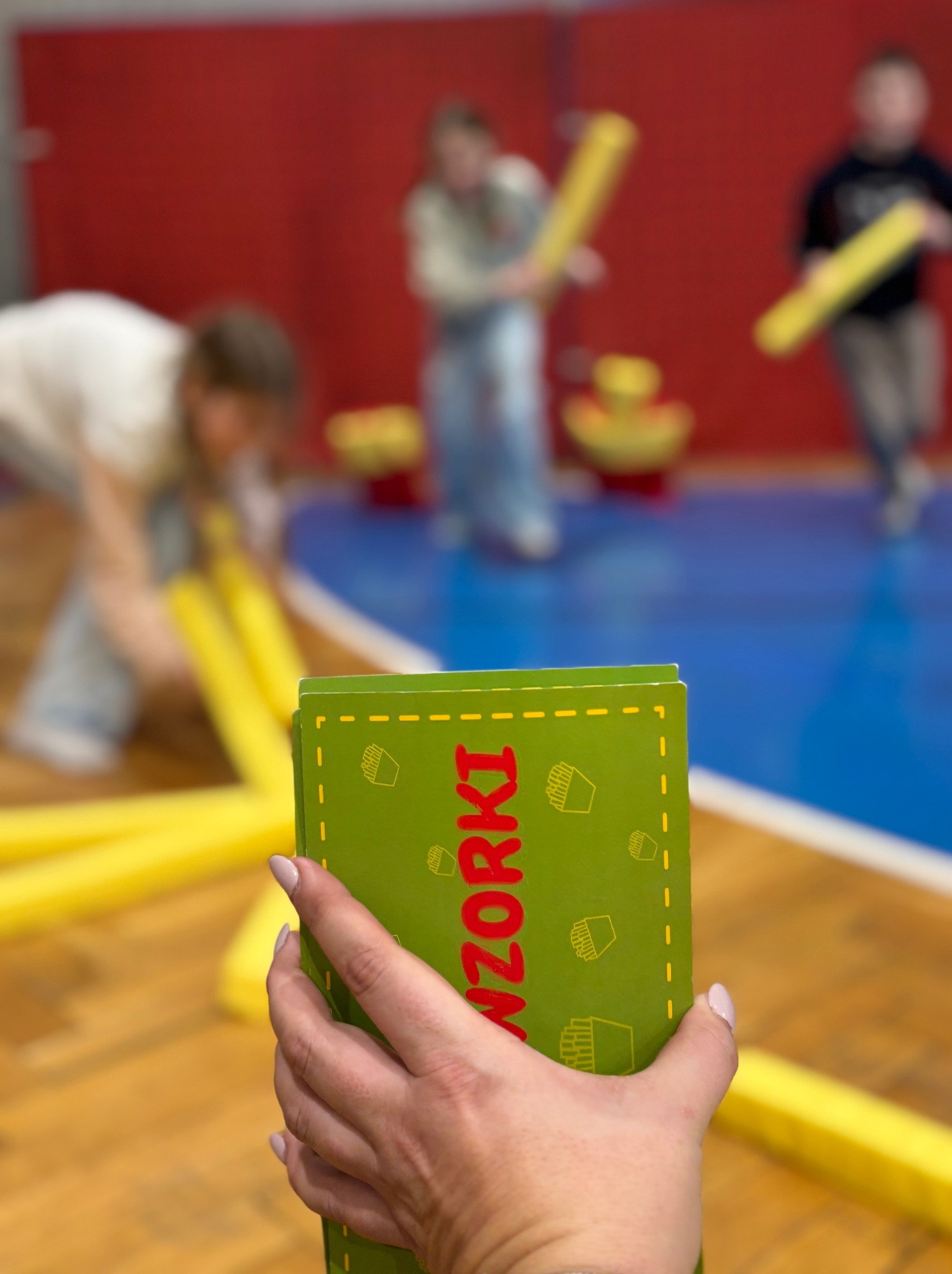 Kobieta trzyma zieloną grę karcianą z napisem 'WZORKI' na tle sali gimnastycznej, gdzie dzieci bawią się z żółtymi piankowymi pałkami.