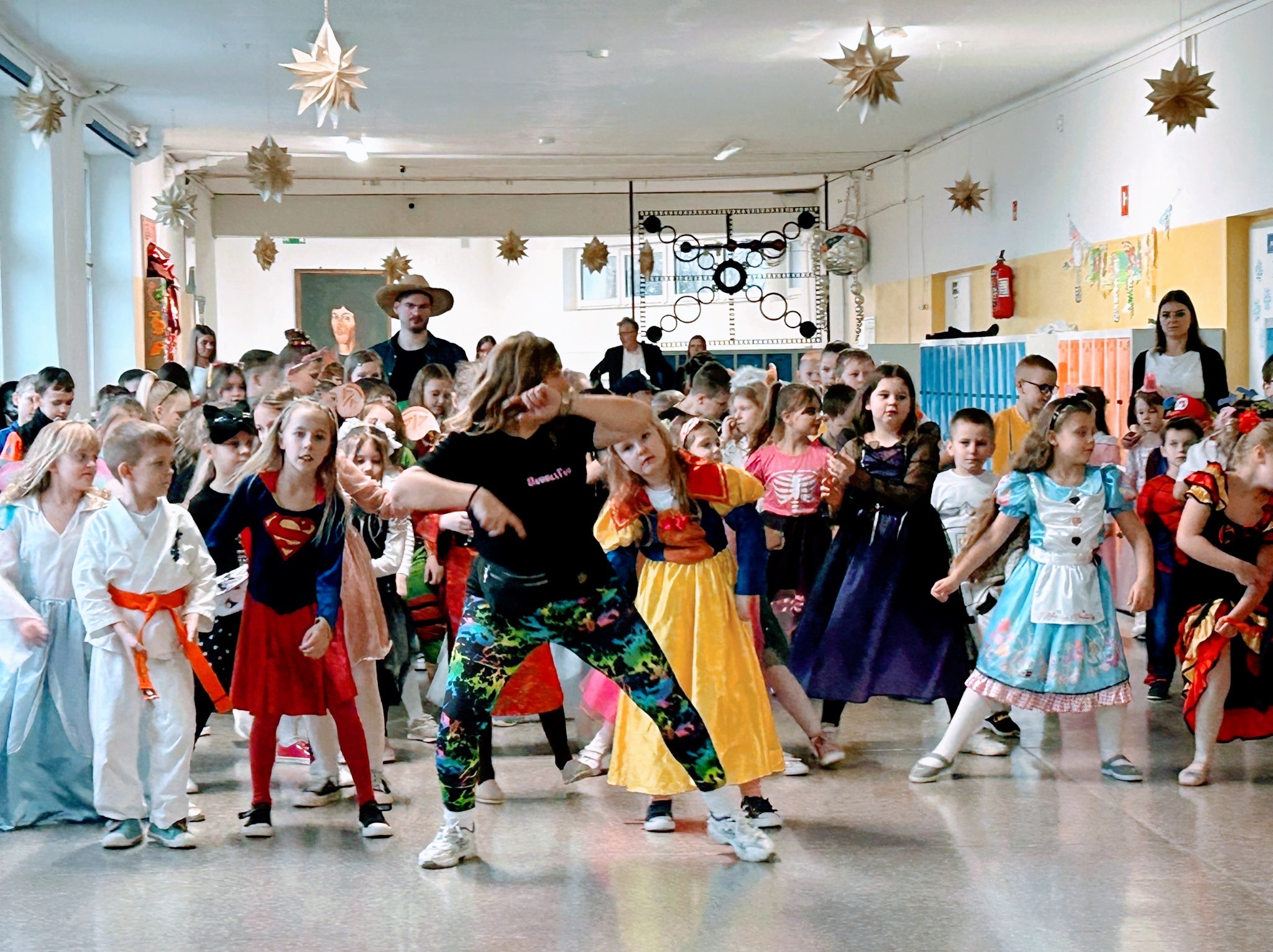 Sala gimnastyczna udekorowana gwiazdami, w której grupa dzieci w kostiumach karnawałowych tańczy pod okiem animatorów, jeden z nich w kapeluszu kowbojskim.