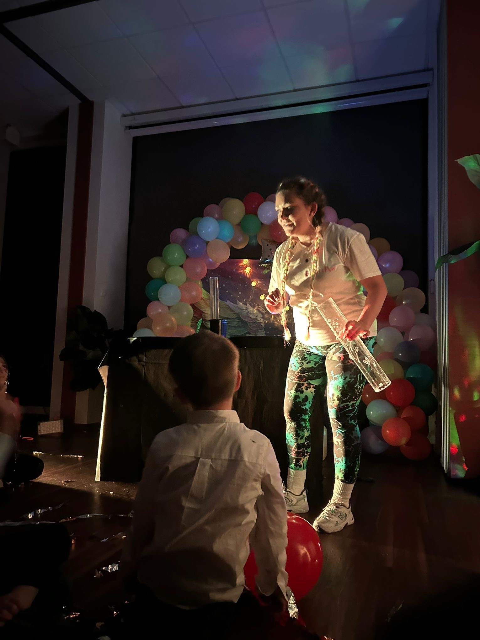 Kobieta w kolorowych legginsach i białej koszulce, z warkoczami, trzymająca szklaną rurkę na tle dekoracji z balonów, podczas występu dla dziecka.