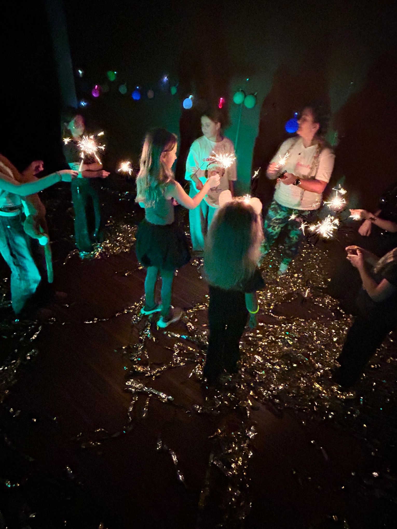 Grupa dzieci i dorośli trzymający zapalone zimne ognie w ciemnym pomieszczeniu udekorowanym balonami i konfetti.