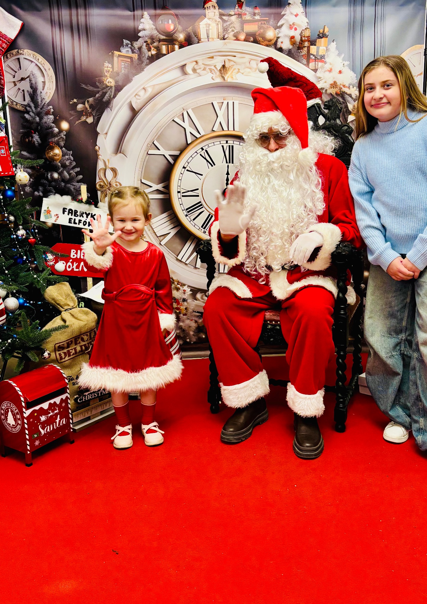 Dwie dziewczynki pozują obok siedzącego Mikołaja na tle dekoracji świątecznych z zegarem i choinką.