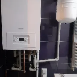 Nowoczesny, biały piec gazowy zamontowany na ciemnych, błyszczących płytkach ściennych, z widocznymi rurami instalacji, manometrem i niewielkim zbiornikiem wyrównawczym.