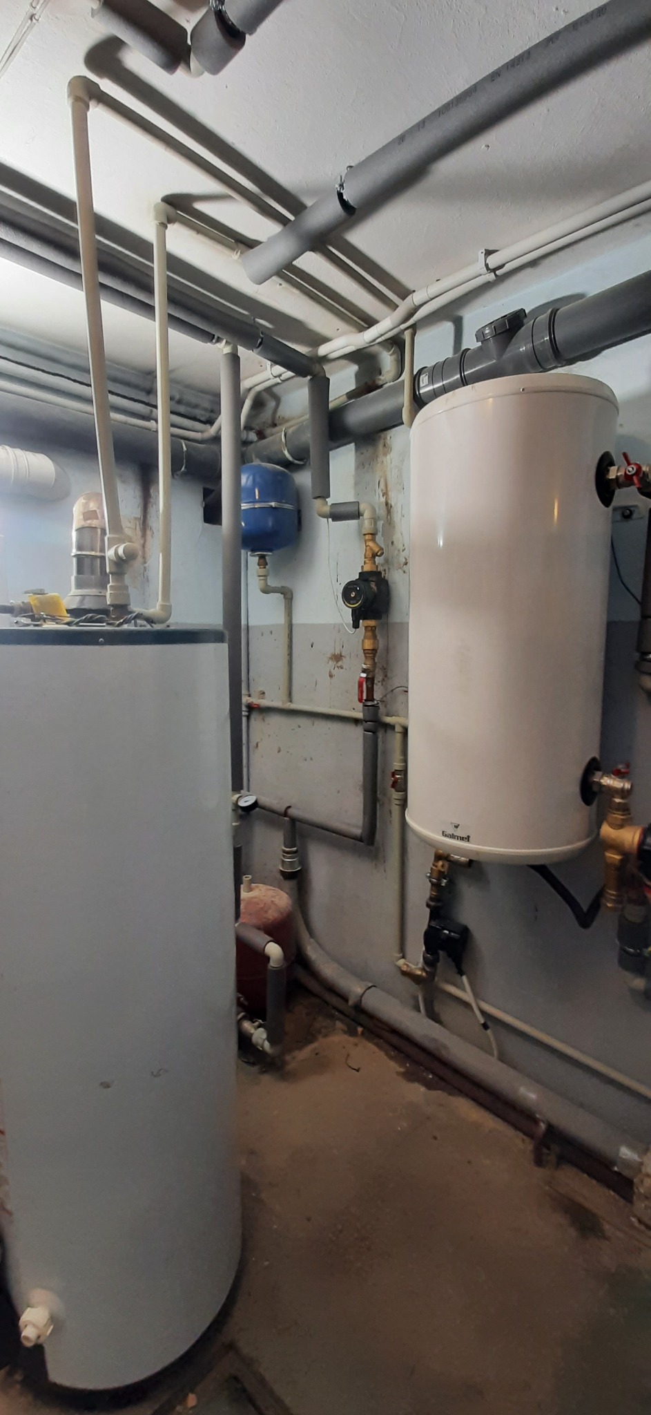 Piwnica z dwoma zbiornikami wody, rurami grzewczymi izolowanymi szarą pianką i zaworami, widoczne ślady wilgoci na ścianach i podłodze.
