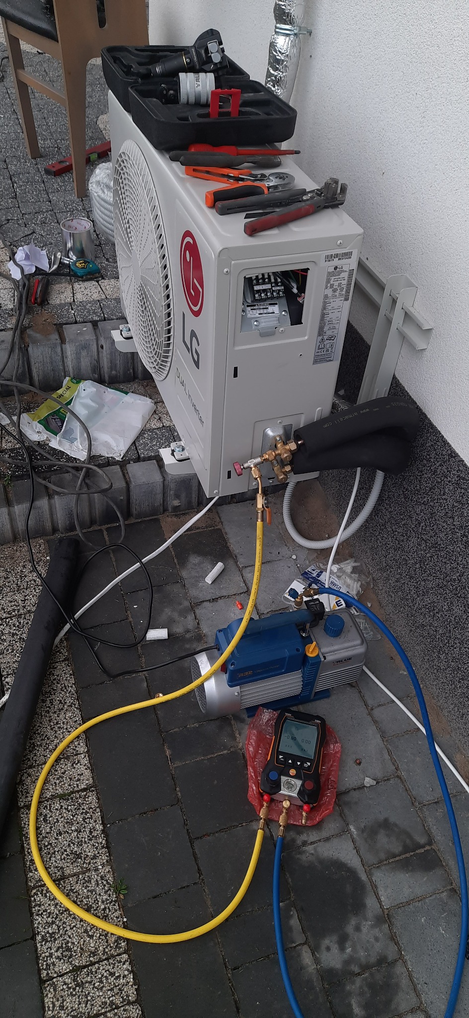 Montaż zewnętrznej jednostki pompy ciepła LG Dual Inverter z podłączoną pompą próżniową i manometrami cyfrowymi, narzędzia w czarnym kufrze na górze jednostki, instalacja na zewnątrz budynku...