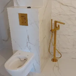 Nowoczesna łazienka z podwieszaną toaletą, złotą armaturą i białymi płytkami imitującymi marmur z geometrycznym wzorem na ścianie.