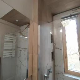 Nowoczesna kabina prysznicowa z deszczownicą i ręcznym prysznicem, wykończona płytkami imitującymi marmur, widoczna przez szklaną taflę i lustro, z drewnianym wykończeniem sufitu i ramy okiennej.