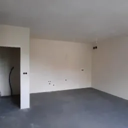 Wnętrze pomieszczenia po tynkowaniu maszynowym ścian i wylewce podłogi, widoczne wyprowadzone kable elektryczne.