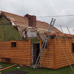 Emil - Budowa domu z bali w trakcie wznoszenia dachu, widoczna więźba dachowa, komin z cegły, drabina oparta o ścianę, folia dachowa i materiały budowlane na trawniku.