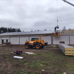 Emil - Montaż paneli dachowych na nowym budynku z białego bloczka; dźwig podnosi panel, a pracownicy na podnośniku go mocują; widoczna konstrukcja więźby dachowej i maszyny budowlane.