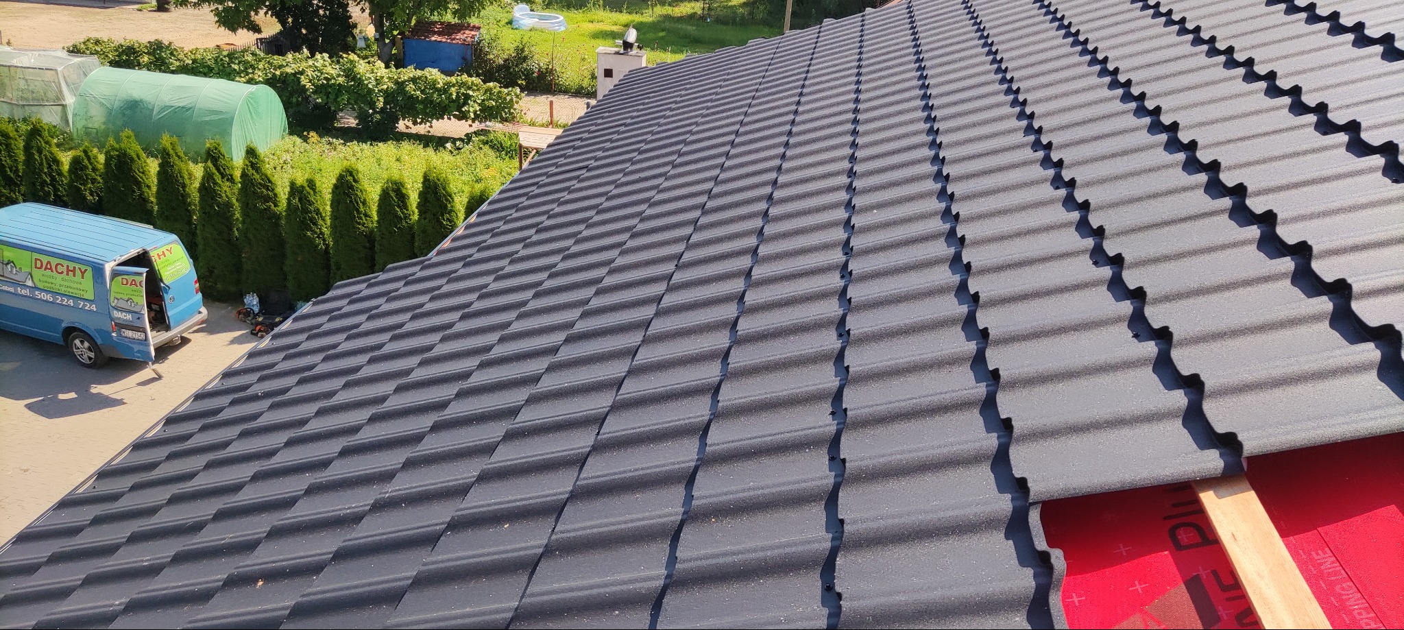Nowy, grafitowy dach z blachodachówki modułowej w trakcie montażu, widoczna membrana dachowa i łaty, na tle zieleni i bus firmy dekarskiej.