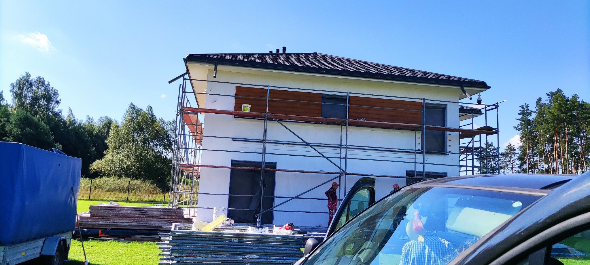 Dwukondygnacyjny dom w trakcie rozbudowy, z zamontowanym rusztowaniem, częściowo wykończoną elewacją z drewnianymi panelami, widocznym sprzętem budowlanym i ekipą pracowników.