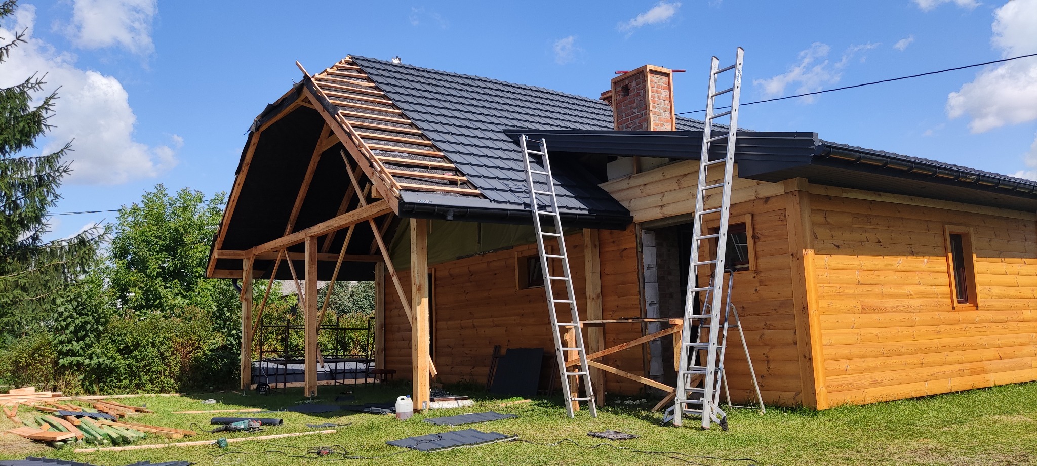 Drewniany dom w trakcie budowy z częściowo pokrytym dachem czarną dachówką, widoczne rusztowanie drewniane, dwie drabiny opierają się o budynek, na trawie leżą deski i narzędzia.
