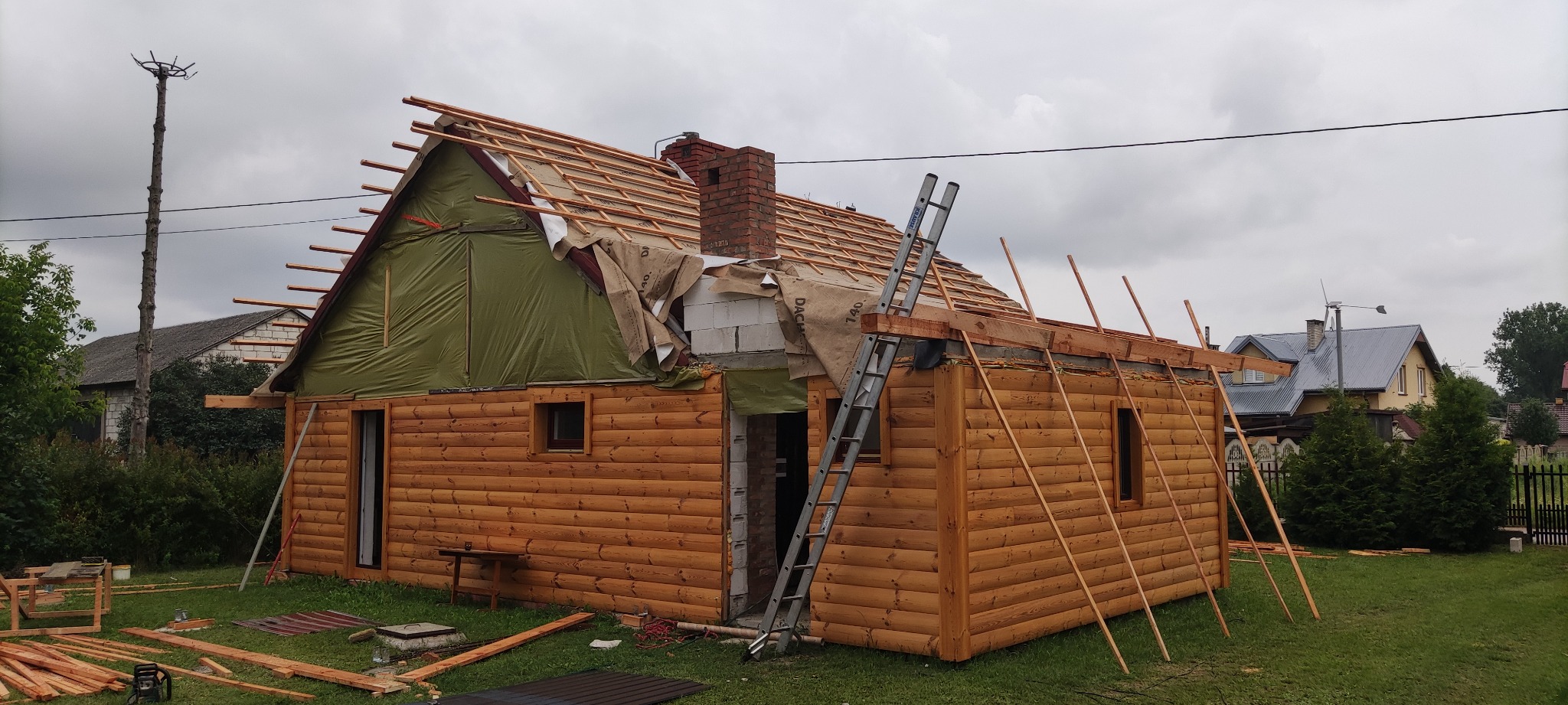 Budowa domu z bali w trakcie wznoszenia dachu, widoczna więźba dachowa, komin z cegły, drabina oparta o ścianę, folia dachowa i materiały budowlane na trawniku.