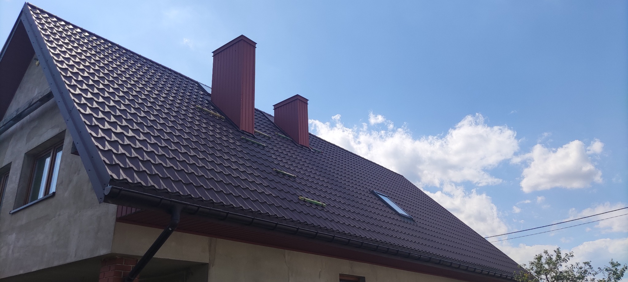 Nowy, brązowy dach z blachy trapezowej z dwoma kominami i oknem dachowym na tle błękitnego nieba z chmurami, Siedlce.