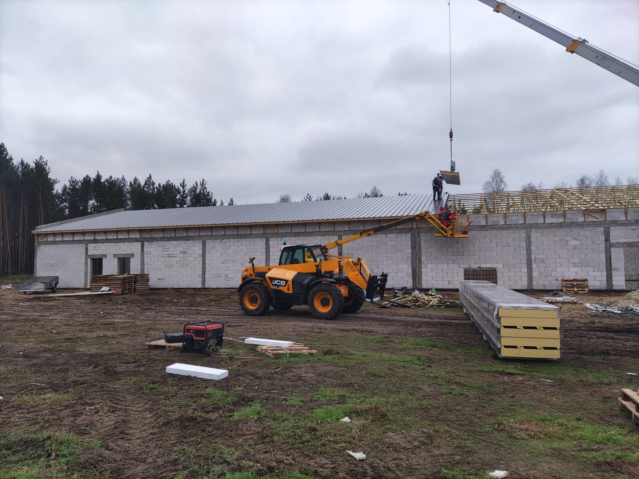 Montaż paneli dachowych na nowym budynku z białego bloczka; dźwig podnosi panel, a pracownicy na podnośniku go mocują; widoczna konstrukcja więźby dachowej i maszyny budowlane.