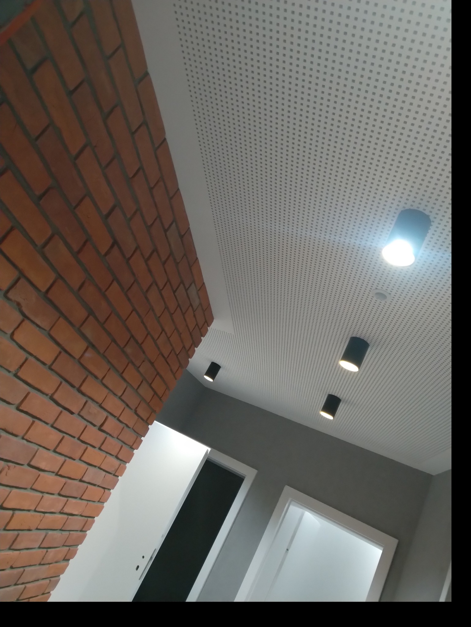 Wnętrze z ceglaną ścianą, perforowanym białym sufitem podwieszanym i czarnymi reflektorami punktowymi, widoczne drzwi w białych ościeżnicach.