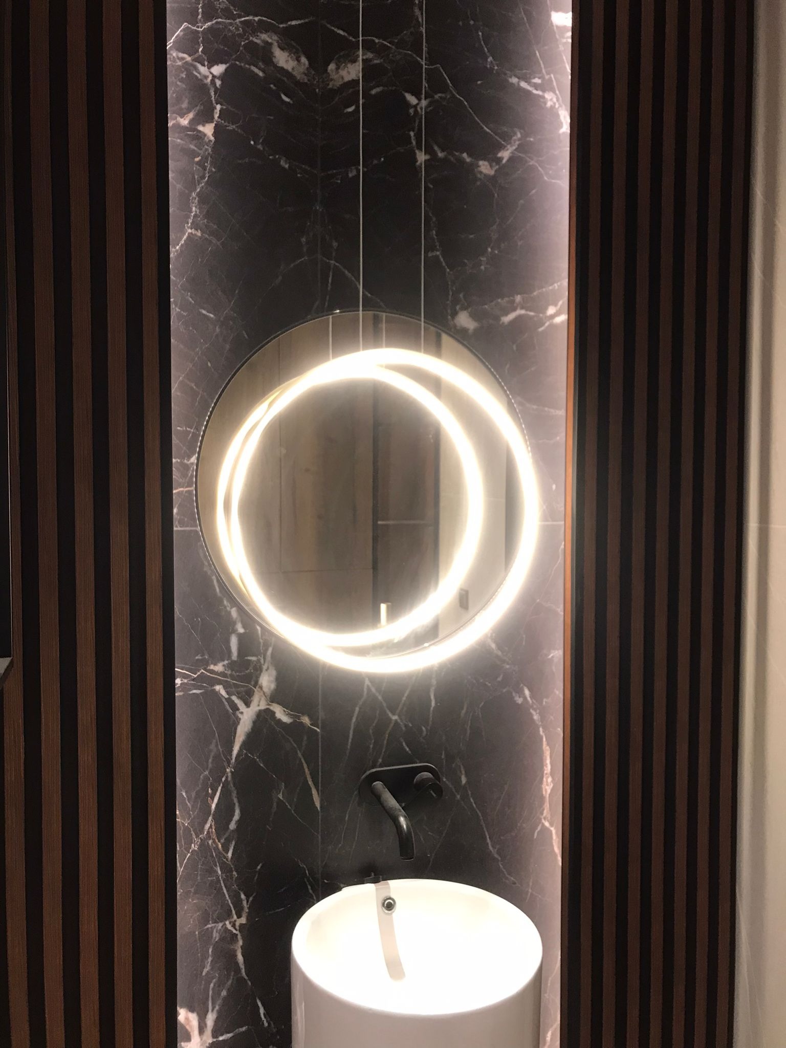 Nowoczesna łazienka z okrągłym lustrem podświetlanym LED, czarną armaturą i panelem ściennym imitującym marmur, uzupełniona pionowymi drewnianymi listwami.
