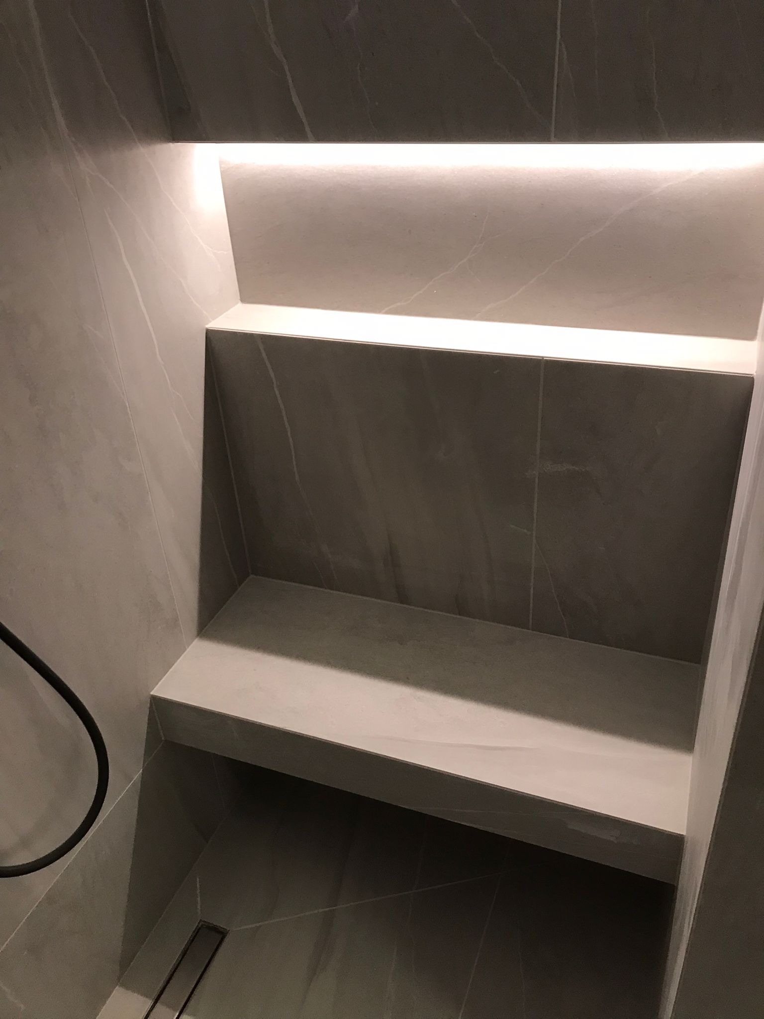 Narożna półka prysznicowa z szarej płyty imitującej marmur, podświetlona od góry ledowym paskiem, z odpływem liniowym w podłodze.